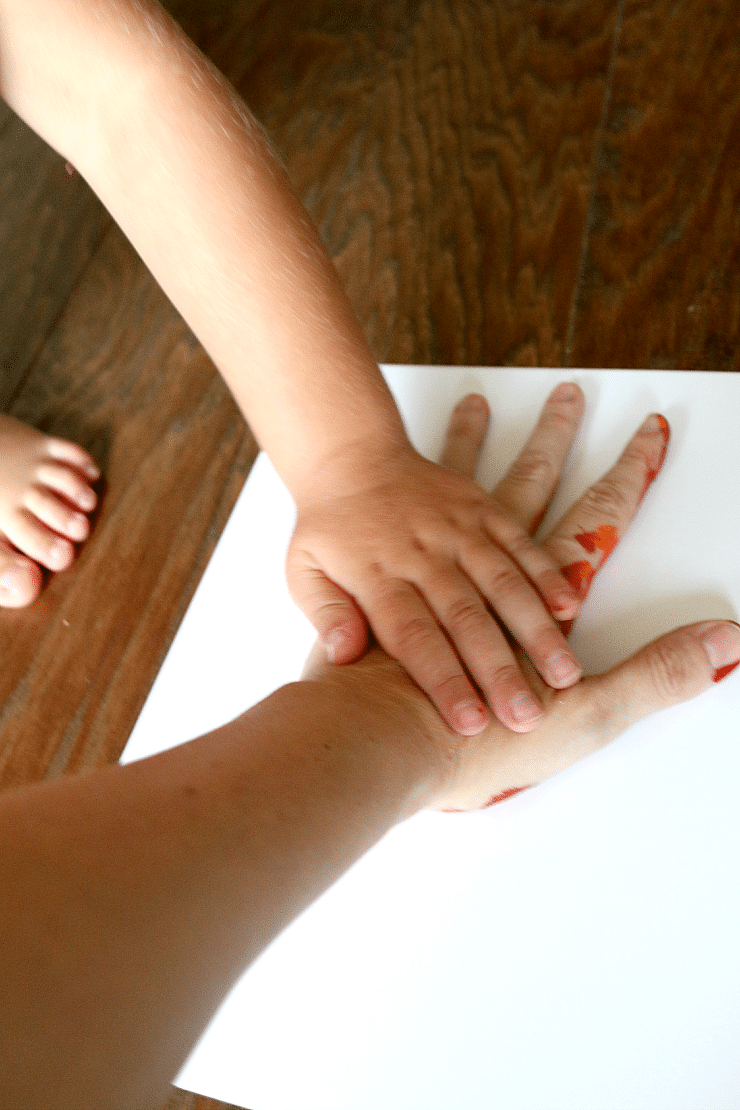 Boy pressing mom's painted hand on a white piece of paper to make a Thanksgiving Gratefulness handprint craft.