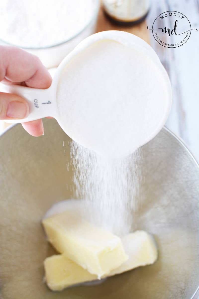 A cup of sugar being poured onto a cup of butter in a mixing bowl to make sugar cookies.