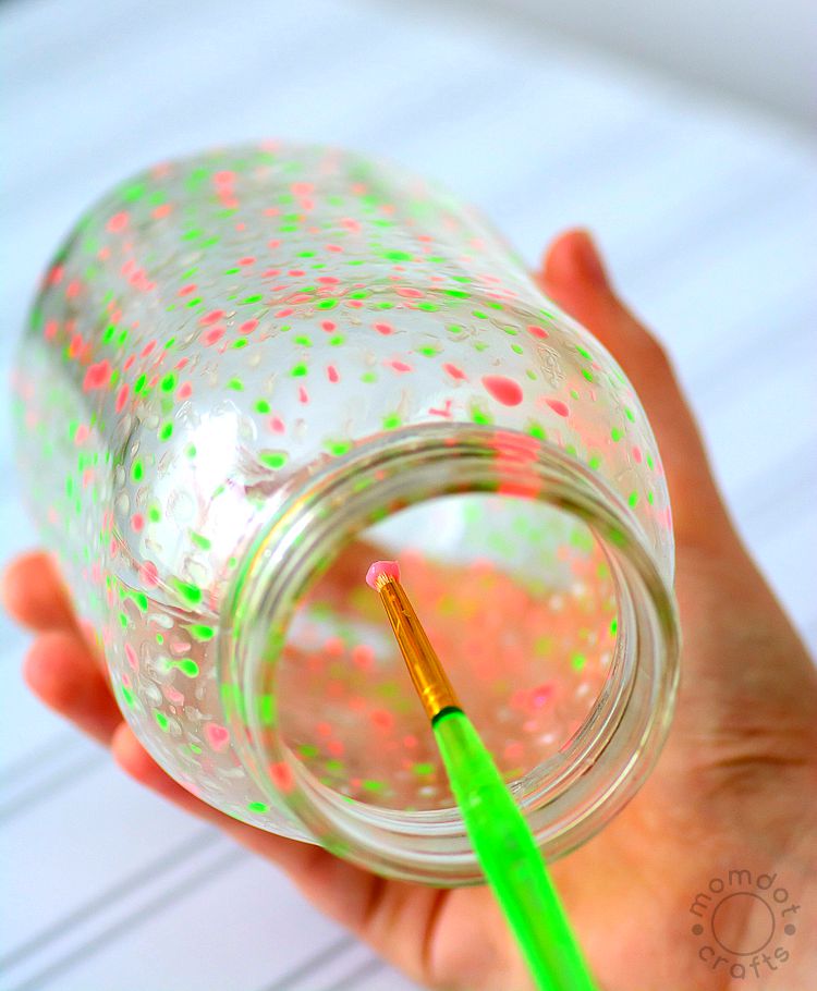 A paintbrush with pink glow-in-the-dark paint inside a mason jar filled with dots of paint