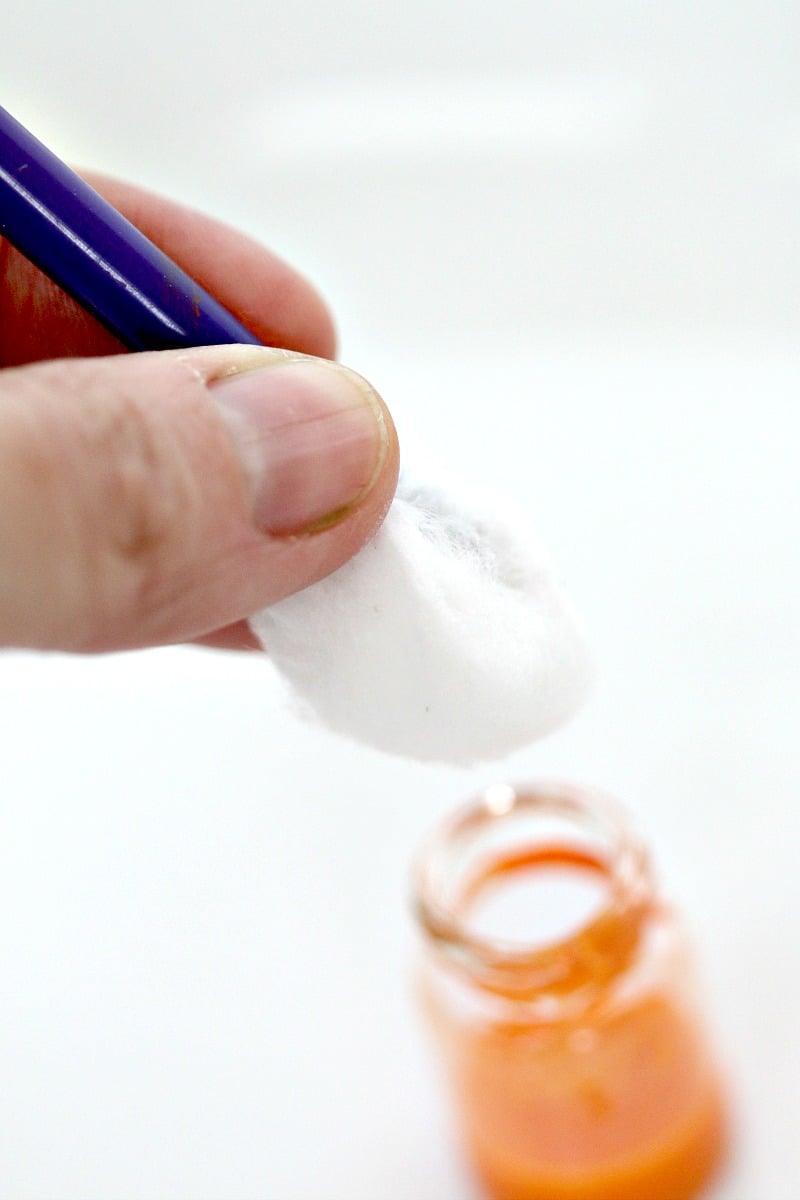 A cotton ball is ready to be stuffed in a small apothecary jar to absorb orange paint and hold it in place inside the jar.