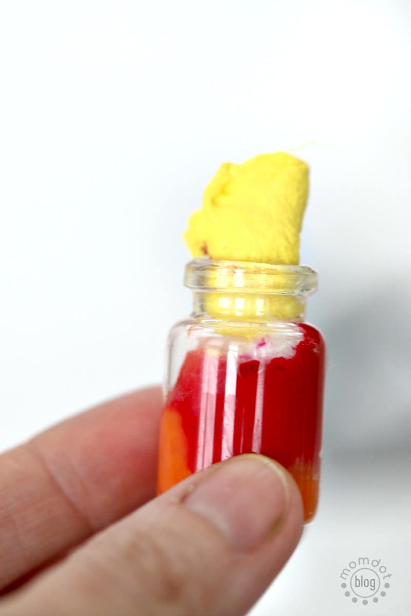 A tiny apothecary jar has an orange cotton ball and red cotton ball inside, and a yellow one being pushed in the top to represent the colors of the sun.