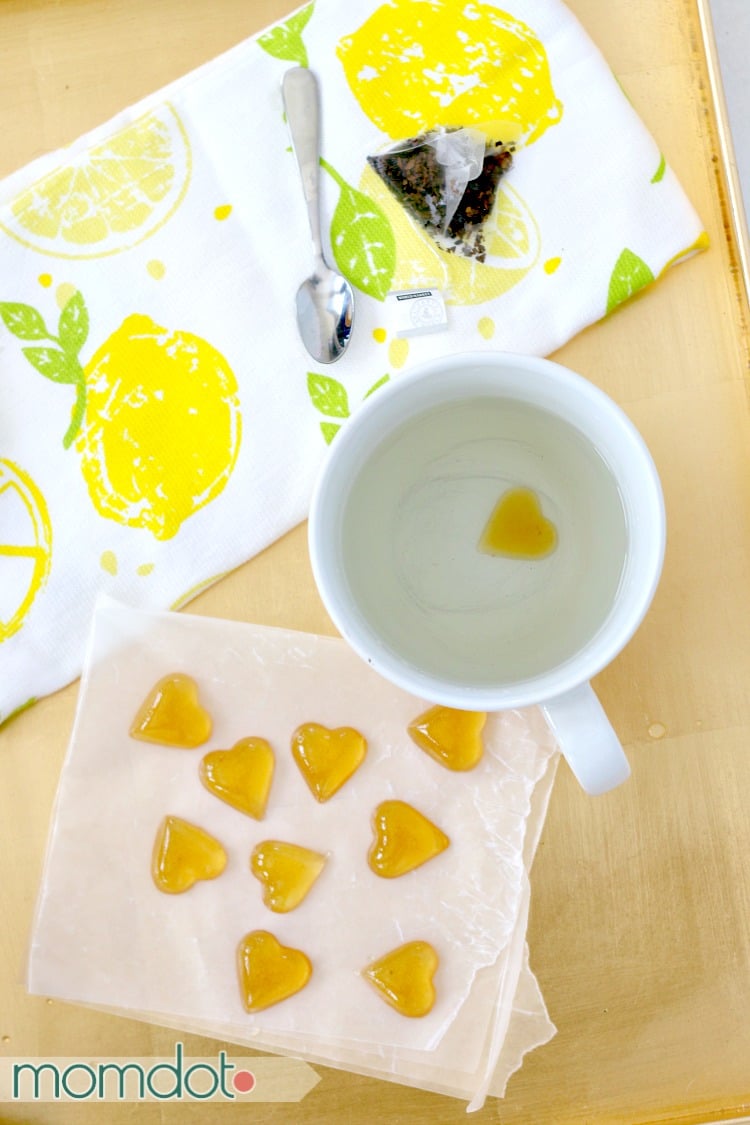 A mug of water with a finished honey drop in the mug and honey drops on waxed paper beside it.