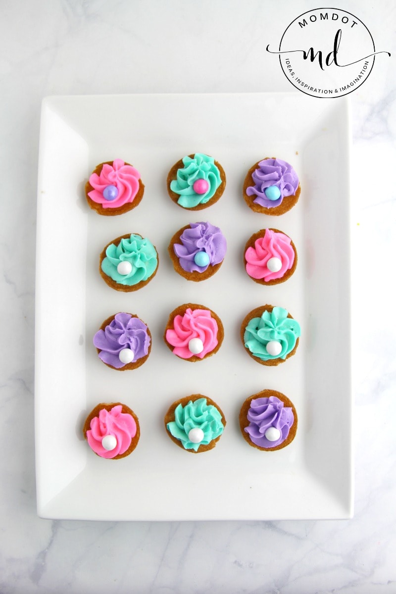 Nilla wafers on a white plate with colored buttercream frosting in the middle and topped with a Sixlet pearl for clam shell cookies
