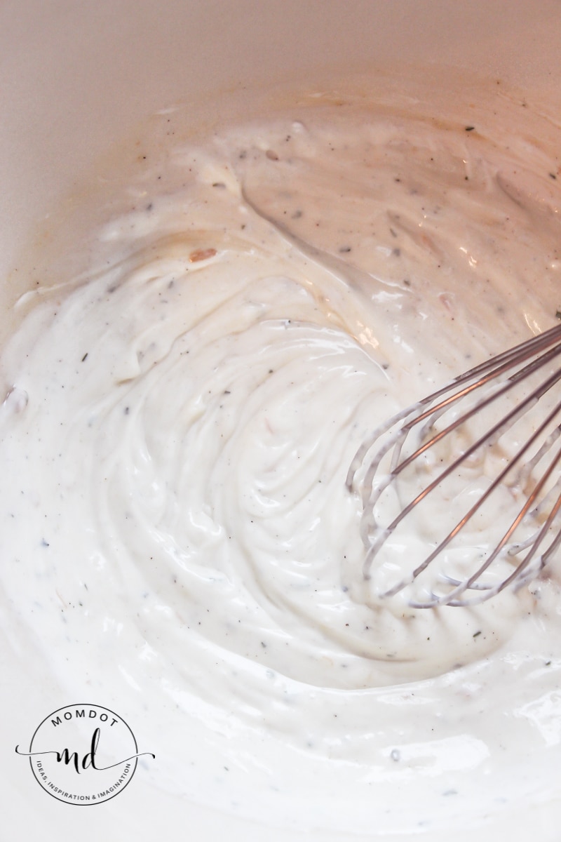 A whisk mixing a bowl of sour cream, cream of chicken soup, and dry ranch dressing powder.