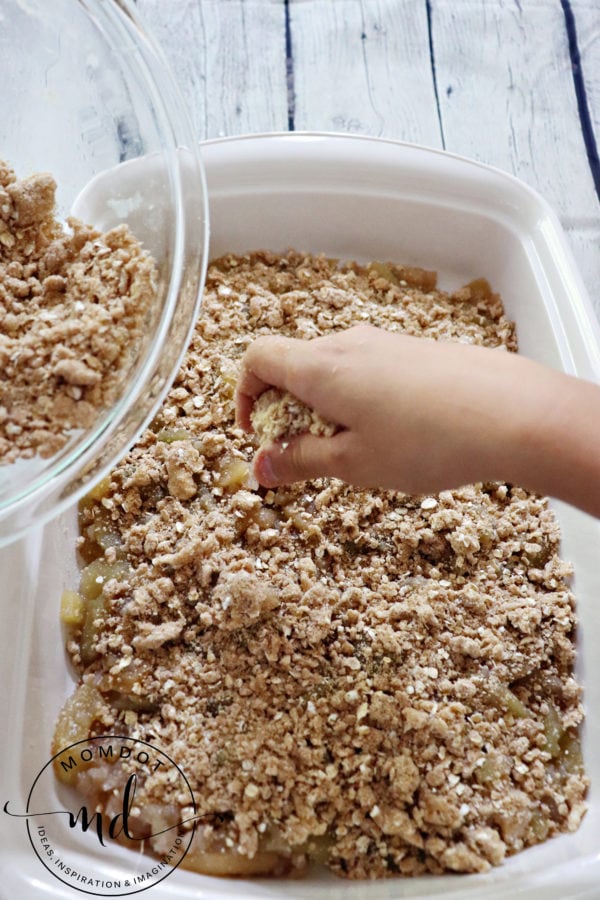 A hand crumbling apple crisp topping over apple pie filling in a casserole dish.