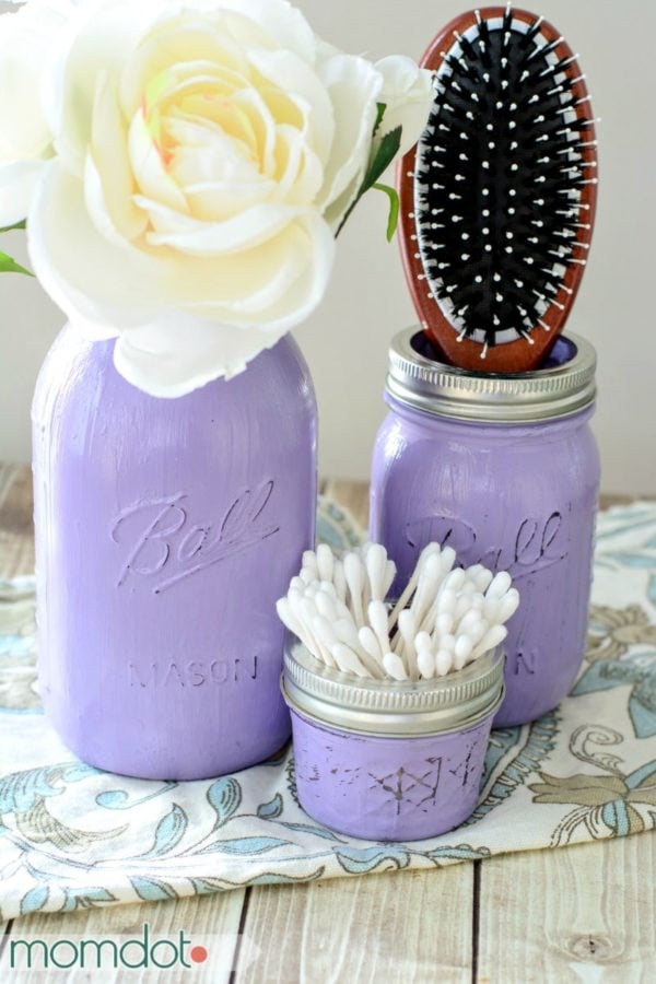 3 purple painted mason jars turned into apothecary jars for the bathroom. One has a rose, one has a hairbrush, and the third is holding Q-tips.